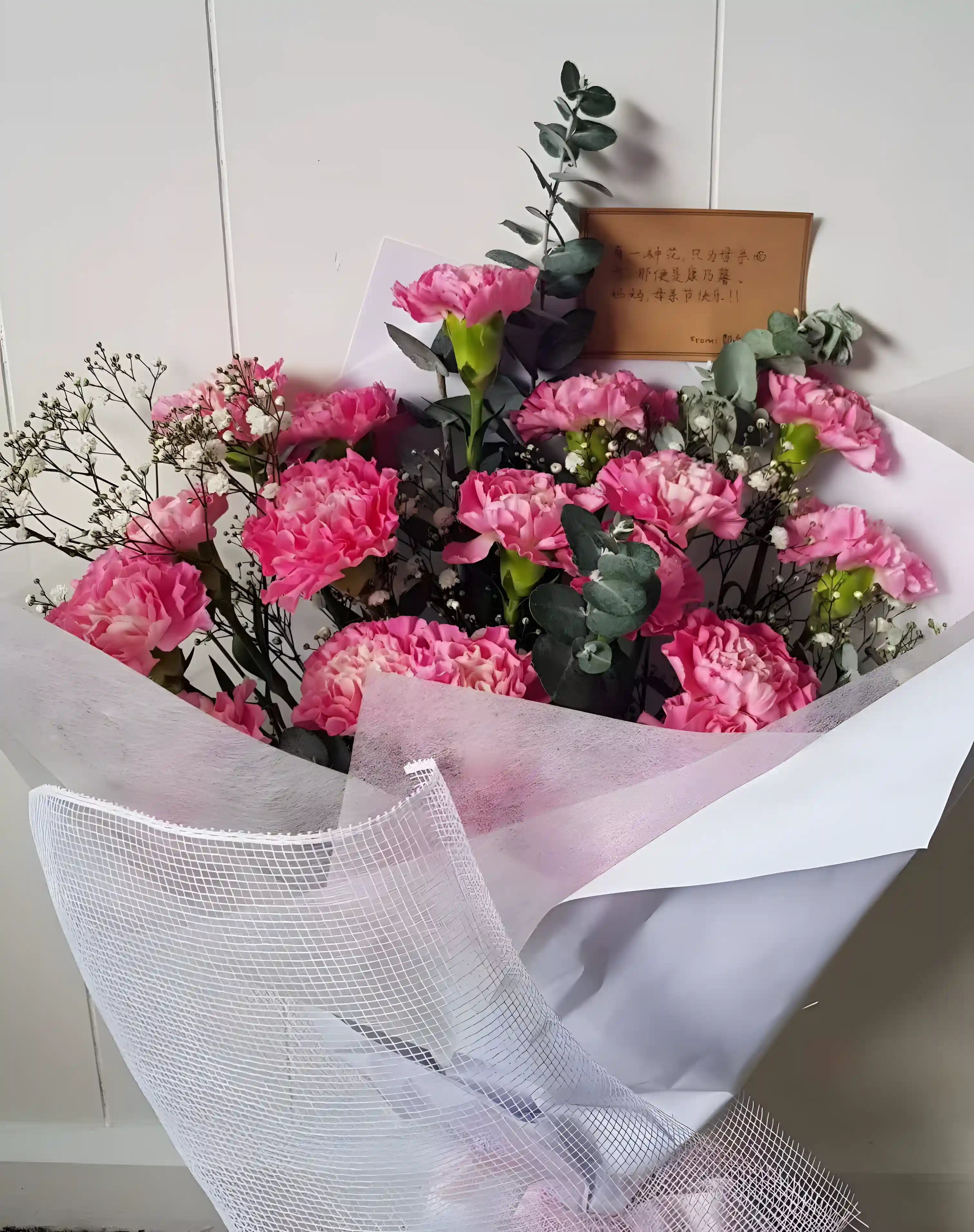A bouquet of pink carnations wrapped in white and pink paper with a brown card and green leaves
